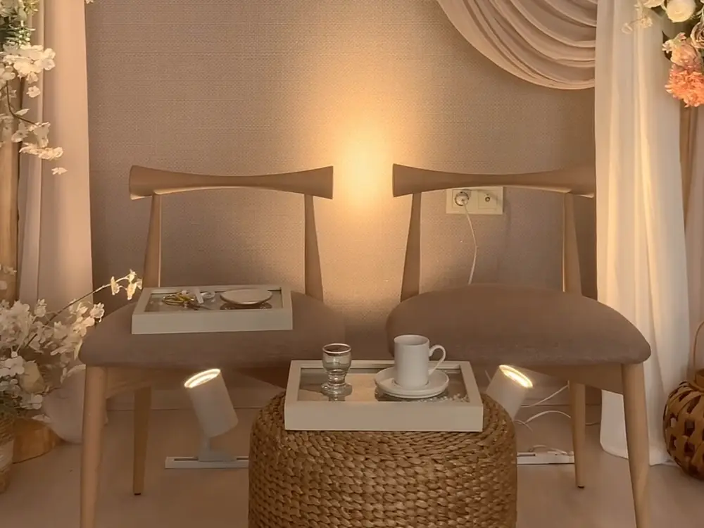 Two wooden chairs in front of a draped cream wall with pink and white flowers, lit by a small spotlight