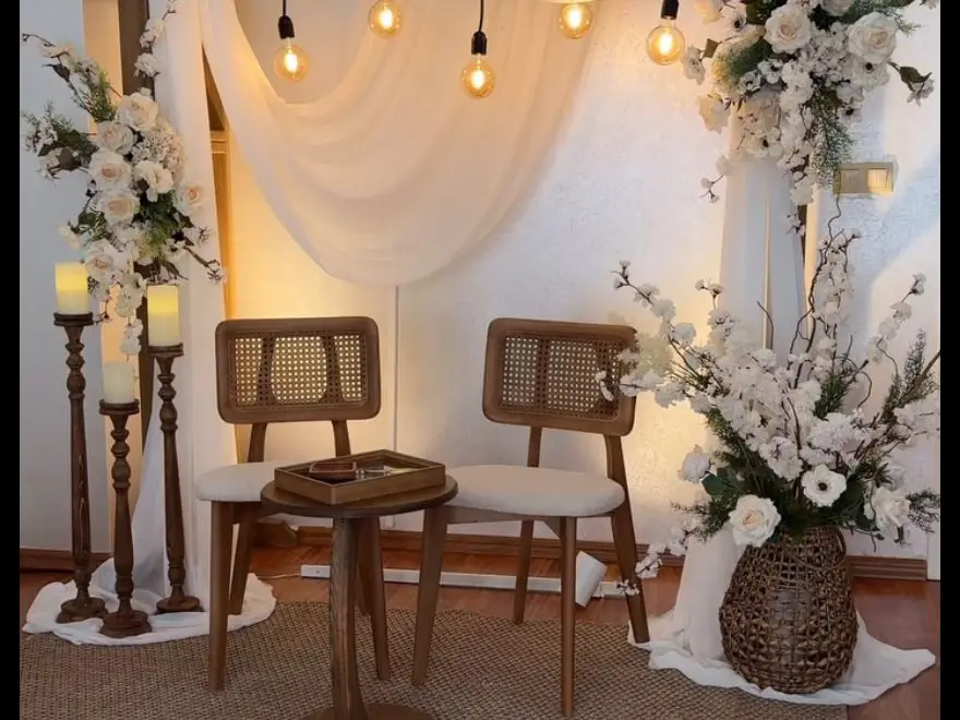 Two cane chairs under a white drape with hanging bulbs, white flowers and candles on tall wooden stands