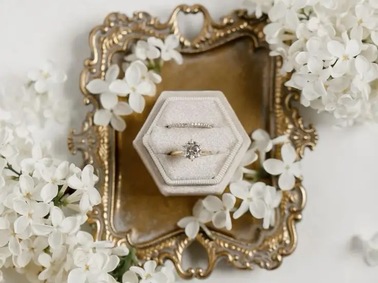 Hexagonal velvet ring box with two rings on a gilded tray surrounded by white lilac blossoms