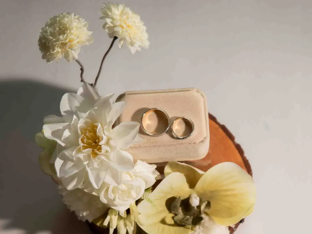 Two wedding bands on a beige ring box with cream dahlia and a pale orchid on a wood slice