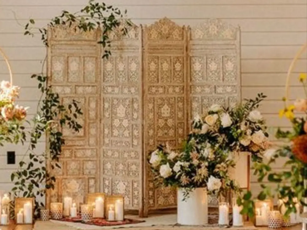 Carved ivory wooden screens with greenery, white roses and candles on a wooden floor against a white wall