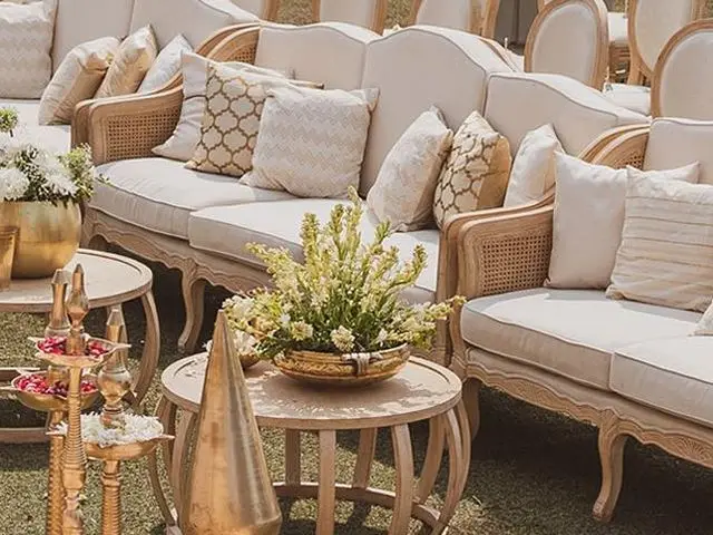 Cream cane sofas with patterned cushions and round wooden tables with brass lamps and flowers on a lawn