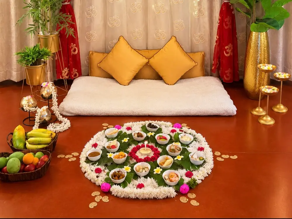 Floor seat with gold cushions, a petal rangoli of bowls and fruit baskets, and a red and cream curtain backdrop