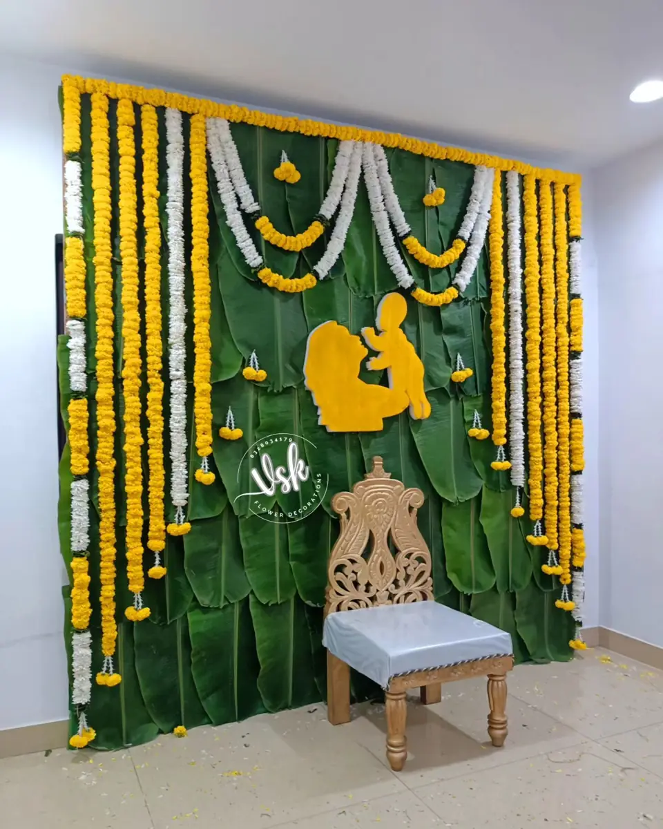 Banana leaf wall with yellow and white marigold strings, a carved chair and a baby motif