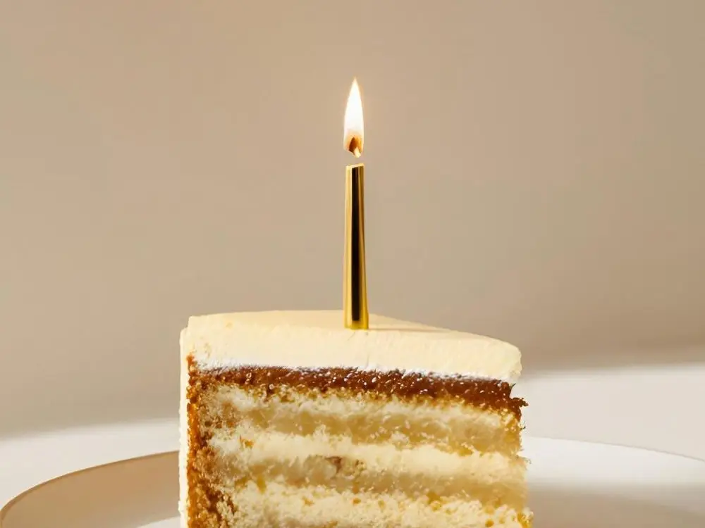 Slice of vanilla cake with a gold candle on a gold-rimmed plate by a cream wall
