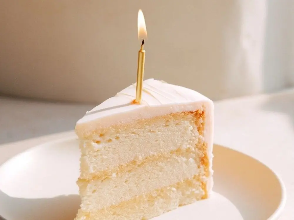 Slice of layered white cake with a lit gold candle on a cream plate