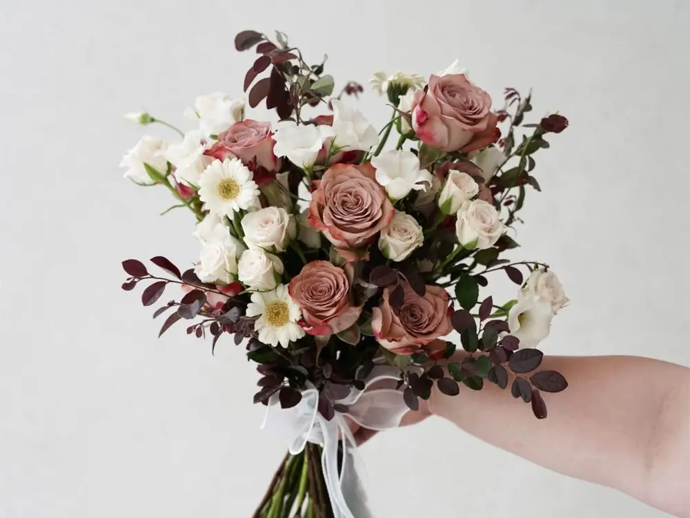 Hand holding a hand-tied bouquet of dusty roses, white daisies and burgundy leaves against a pale wall