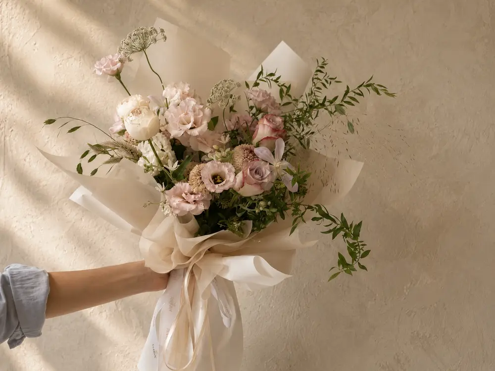 Pastel bouquet wrapped in cream paper and ribbon held up against a sunlit beige wall