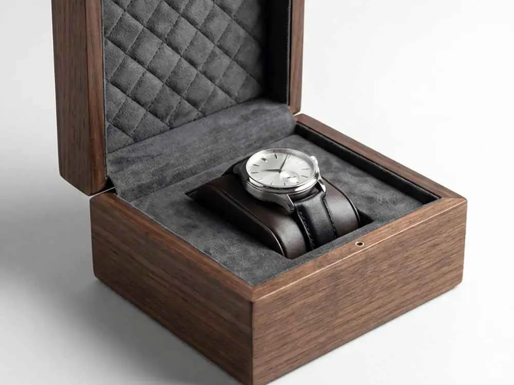 Open walnut watch box with a steel watch resting on a grey quilted cushion