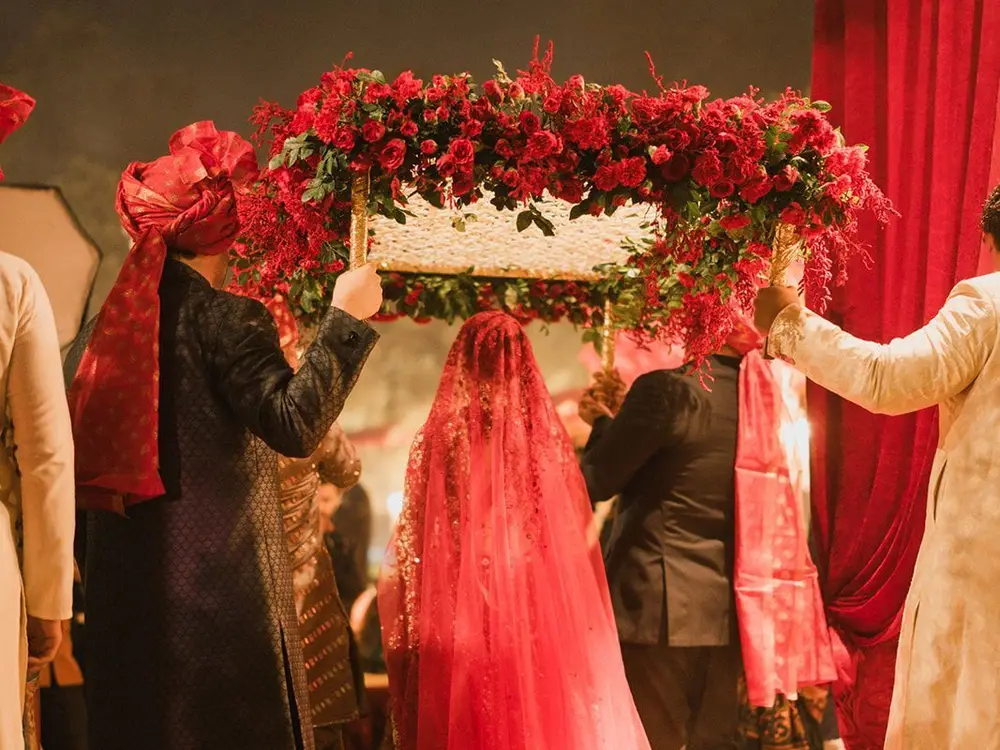 Bride seen from behind in a red veil walking under a red rose canopy carried by relatives at dusk