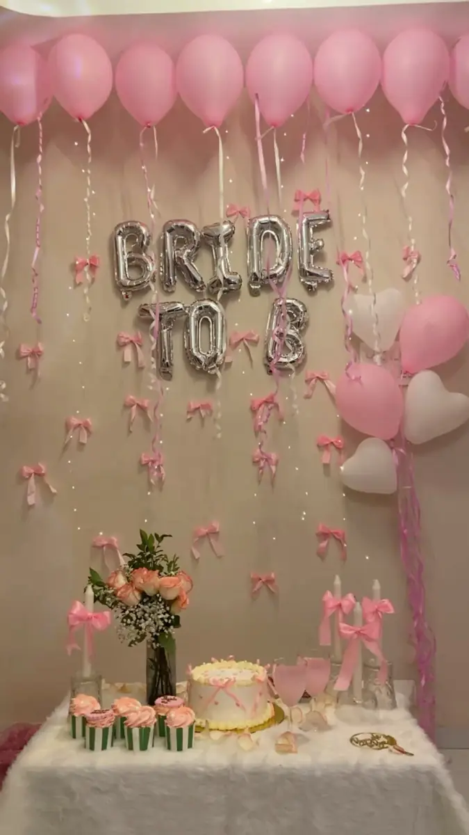 Silver BRIDE TO B foil letters under pink ceiling balloons with a cake table and pink roses