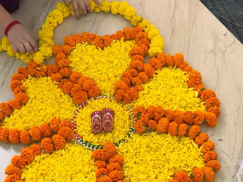 Hands finishing a flower rangoli of yellow and orange marigold petals with a marigold garland outline on a marble floor