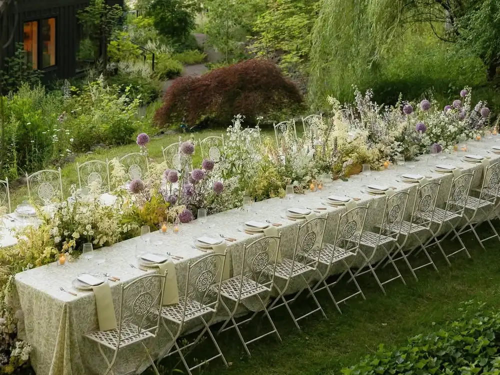 Long garden table laid with white linen, small vases of wildflowers and folding chairs on a lawn