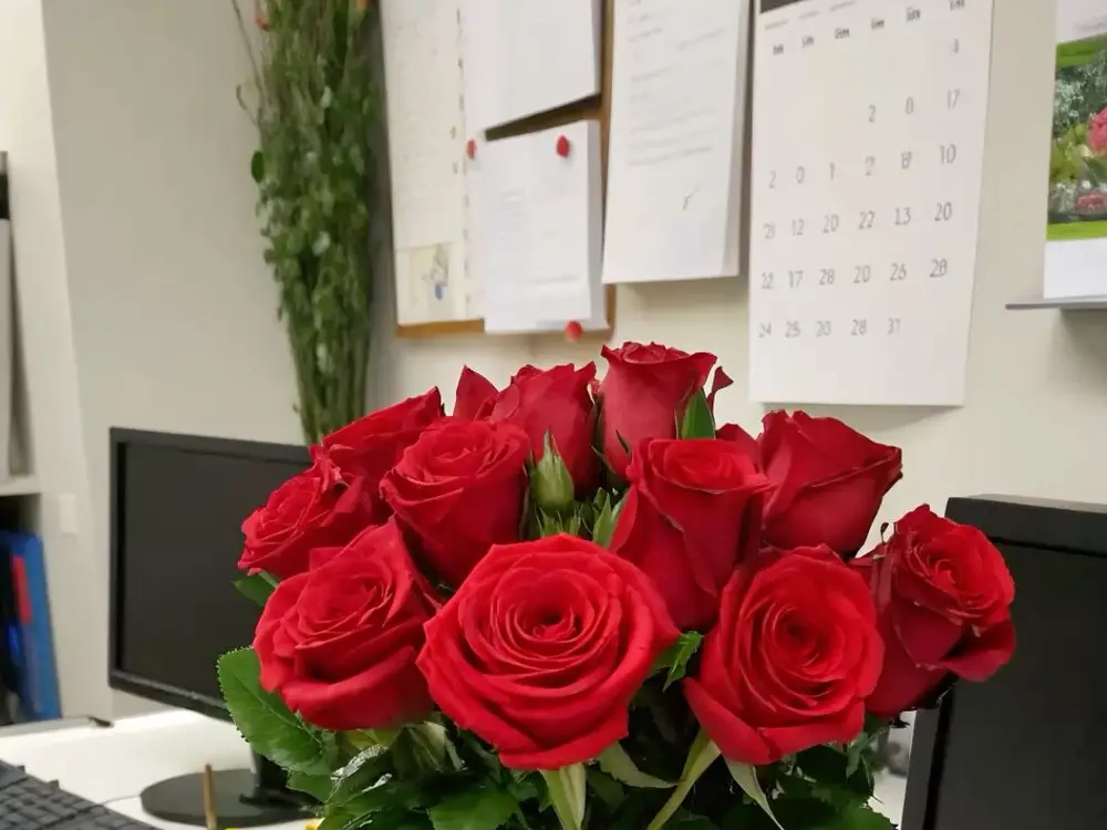 Red roses in a glass vase on a white office desk beside a monitor and a wall calendar