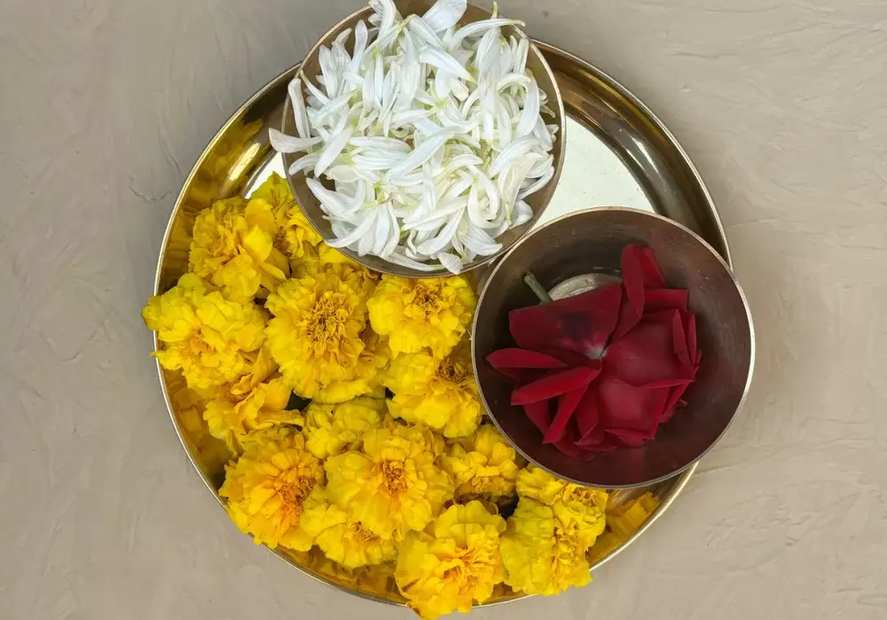 Brass thali with yellow marigold, a bowl of white jasmine petals and a bowl of red rose petals on a beige surface