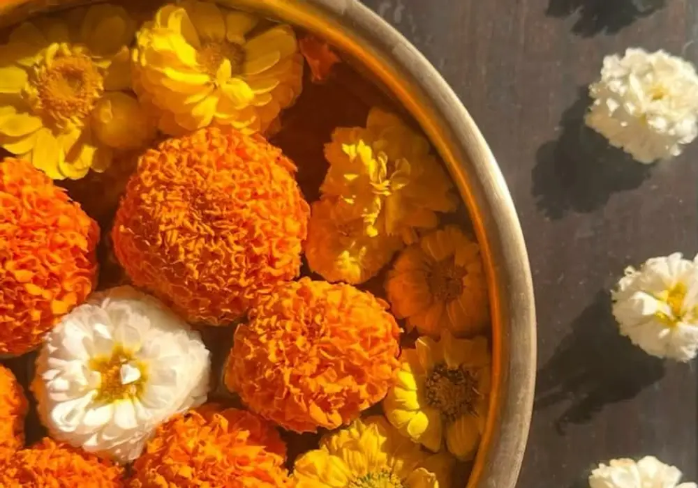 Orange marigold and yellow chrysanthemum heads floating in a brass bowl with white flowers on a dark wooden table