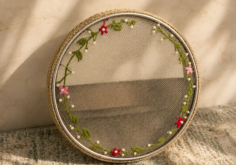 Round sieve with a hand-embroidered wreath of small pink and red flowers resting against a cream wall in soft light