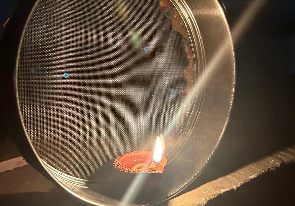 A steel sieve with a single lit clay diya inside it in the evening light
