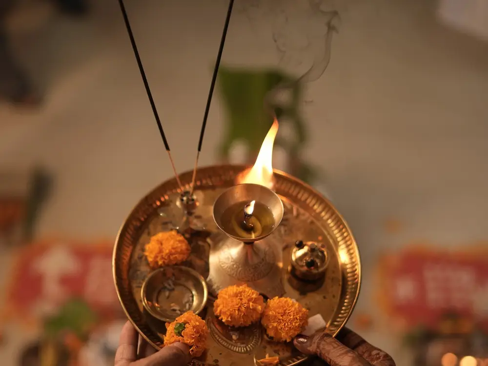 Hands holding a brass aarti thali with a lit diya, incense sticks and three marigold flowers