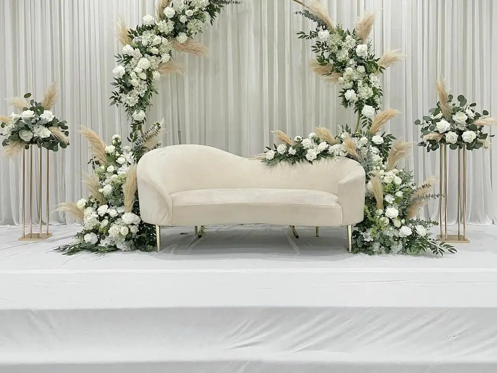 Gold ring arch with white roses, greenery and pampas grass behind a cream sofa on a white platform