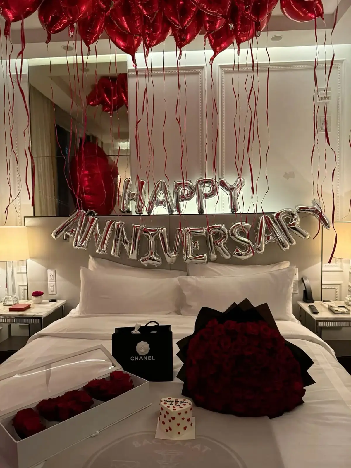 A hotel bed with red heart balloons, Happy Anniversary foil letters and a rose bouquet