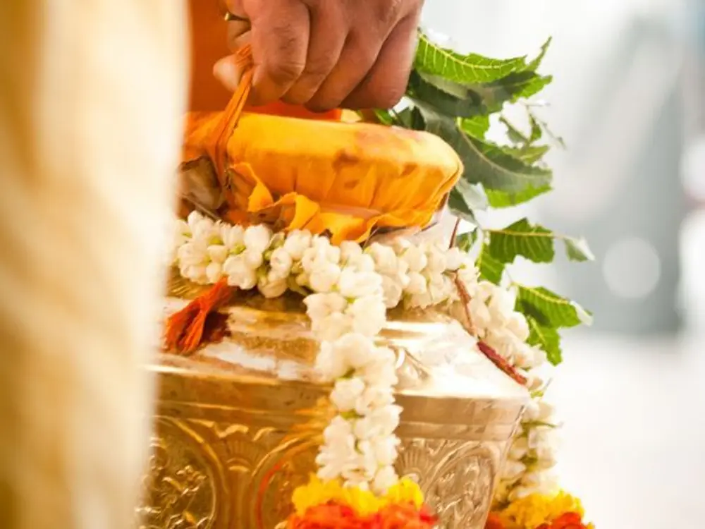 Pandit's hand holding a kalash with mango leaves, coconut and a jasmine garland