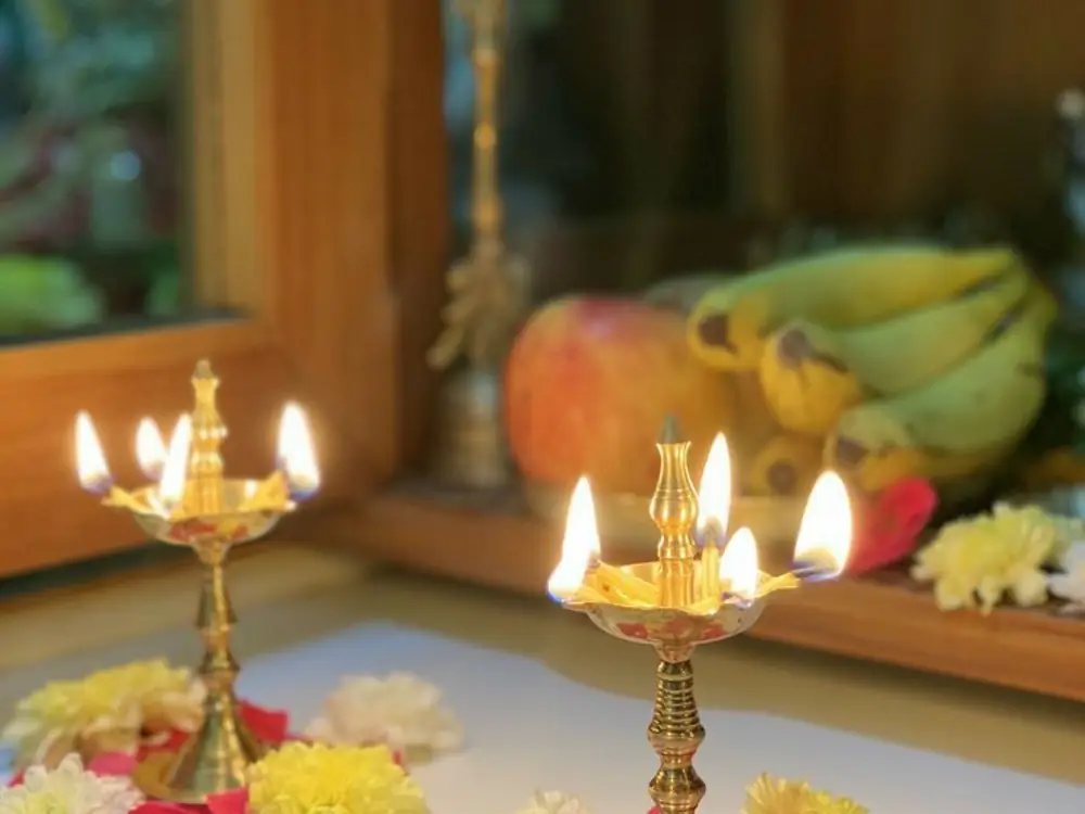 Two brass lamps burning beside fruit and yellow flowers on a puja shelf