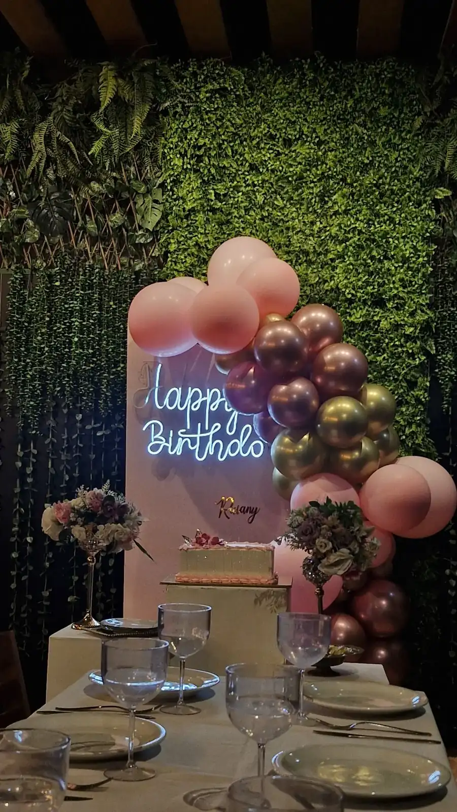 Birthday corner setup at a cafe with balloon garland on a green hedge wall and cake stand