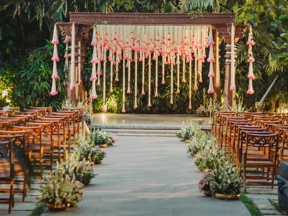 Wooden mandap in a garden with hanging white and pink flower strings and brass lamps, with chairs set in rows facing it