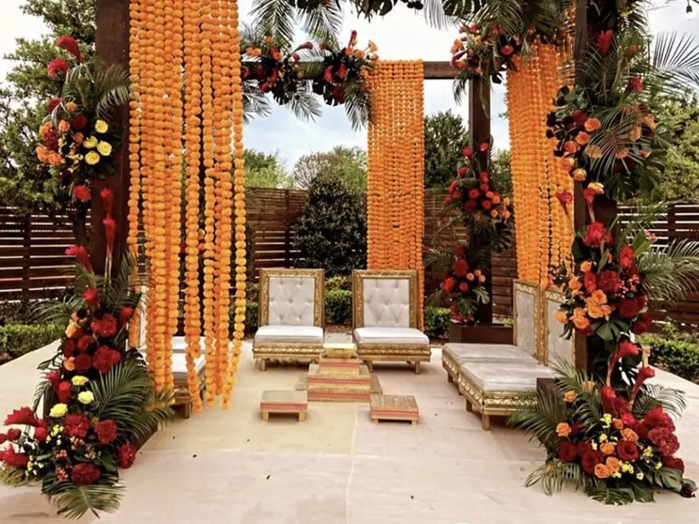 Mandap with orange marigold curtains hanging between wooden pillars, with red rose and palm leaf arrangements and white seats